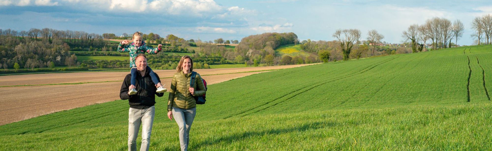 Vader en moeder met kind in de nek wandelen door groen weiland. 